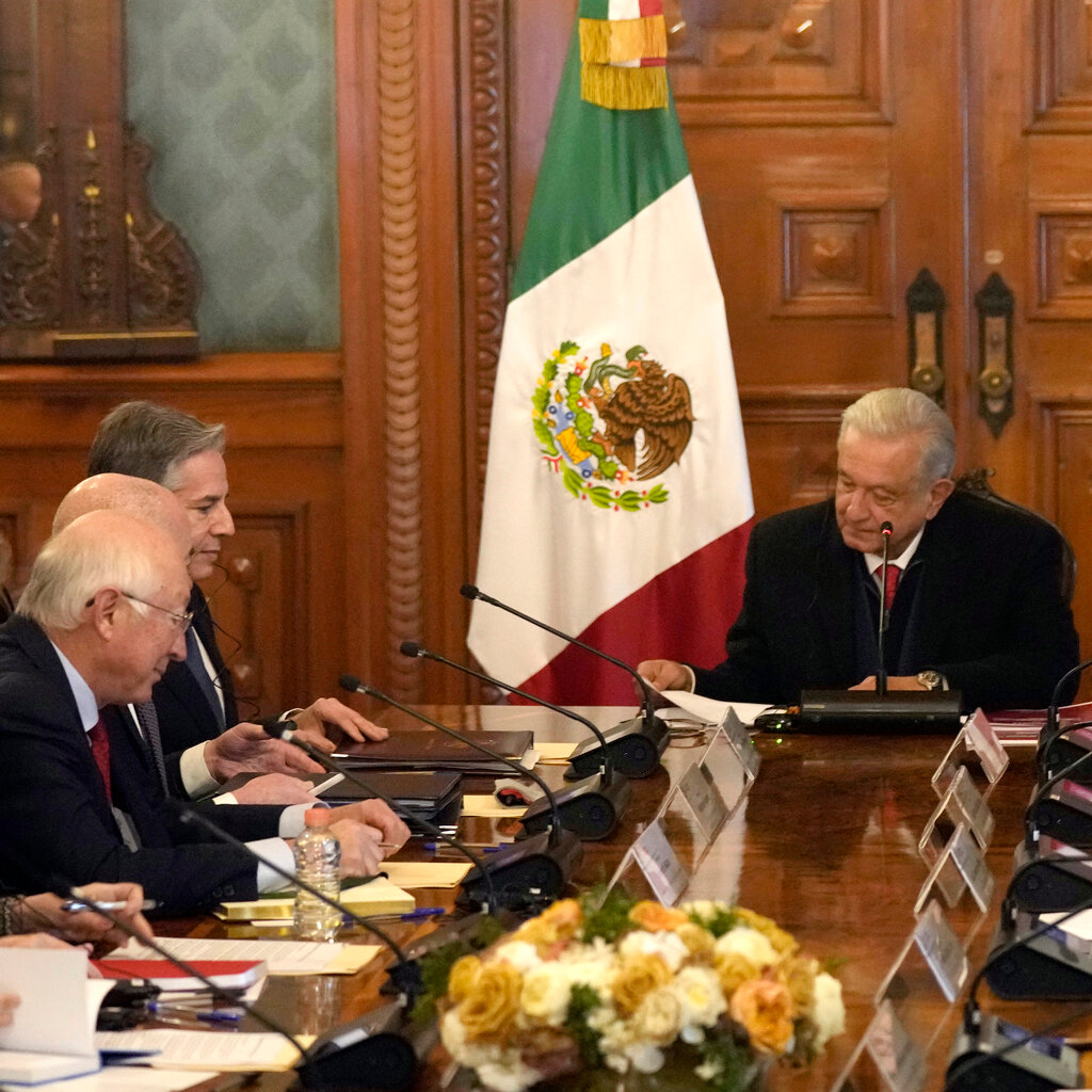 Men sit in a wood paneled room in front of microphones. A Mexican flag is in the background. 