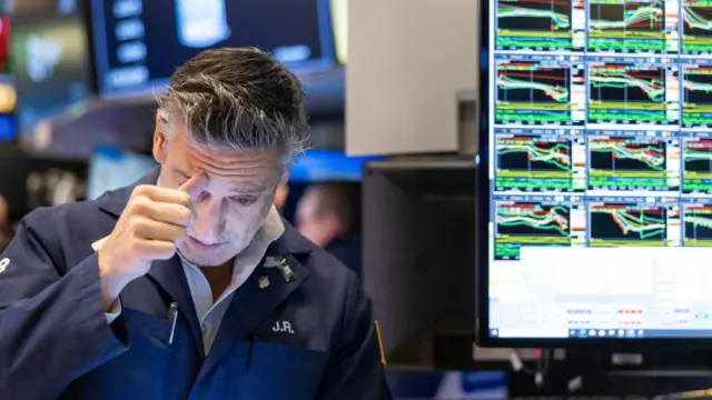 A trader looks concerned at his screen with a screen showing various charts in the background. A trader looks concerned at his screen with a screen showing various charts in the background.